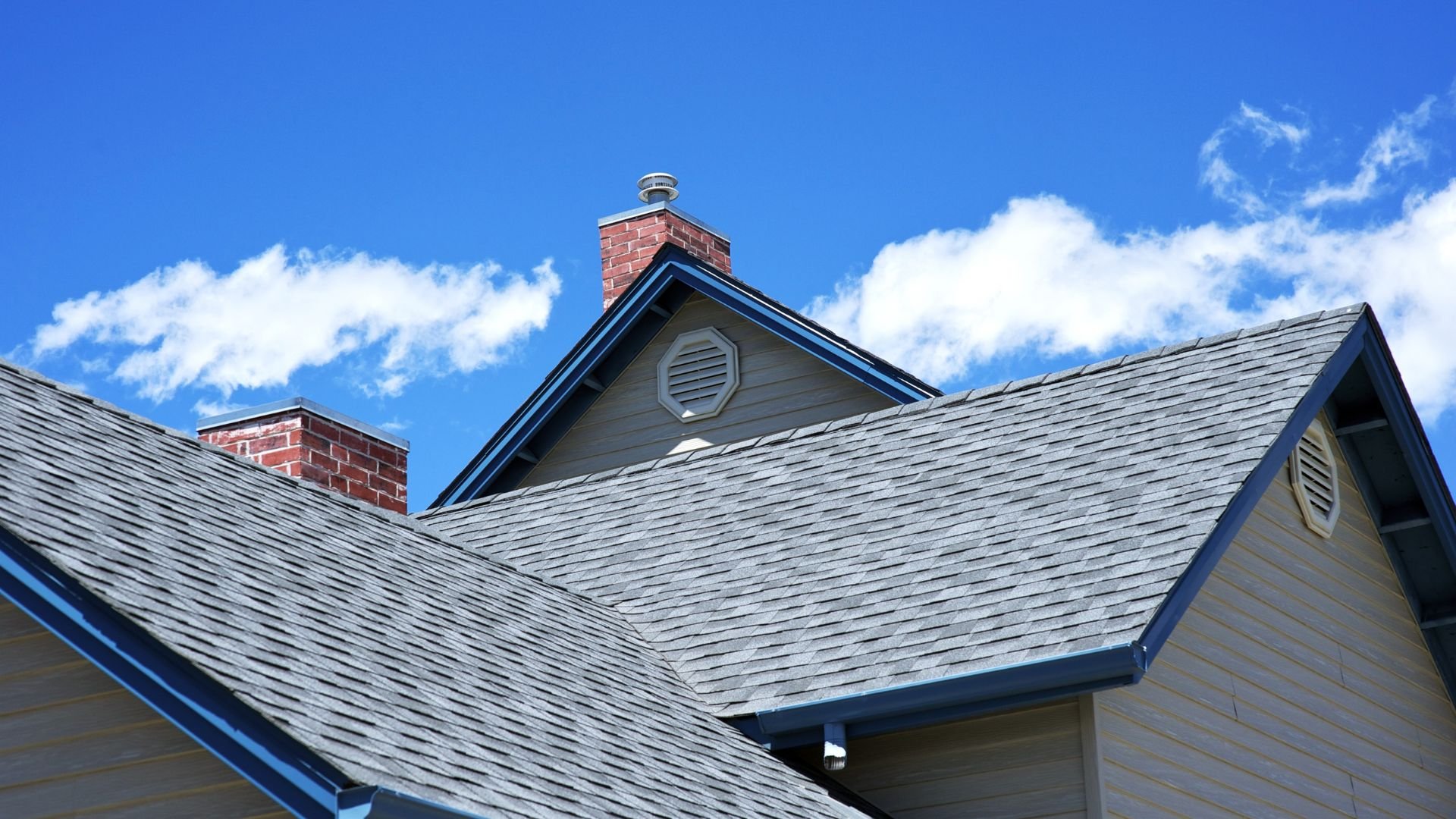 Gray shingle roof with brick chimneys against blue sky with white clouds