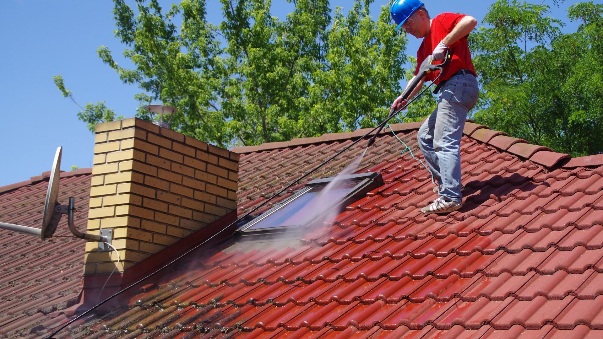 Worker in blue hard hat power washing red tile roof on sunny day